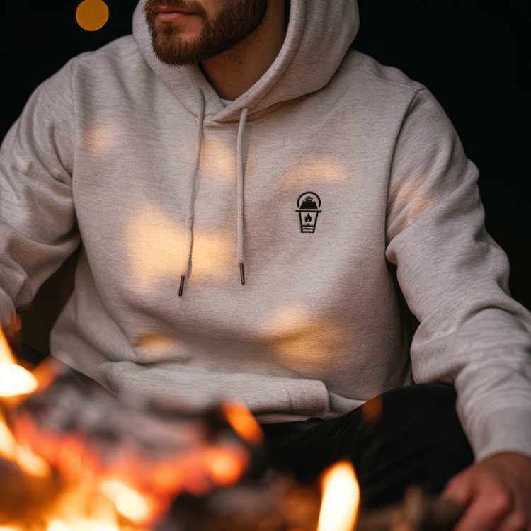 A man wearing a light Gaslamp Ash hoodie with a small black chest graphic, sitting near a fire with warm light reflecting on the fabric.